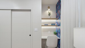 Una habitación moderna y luminosa, con una puerta blanca, un escritorio con silla y una estantería con libros y una planta.
