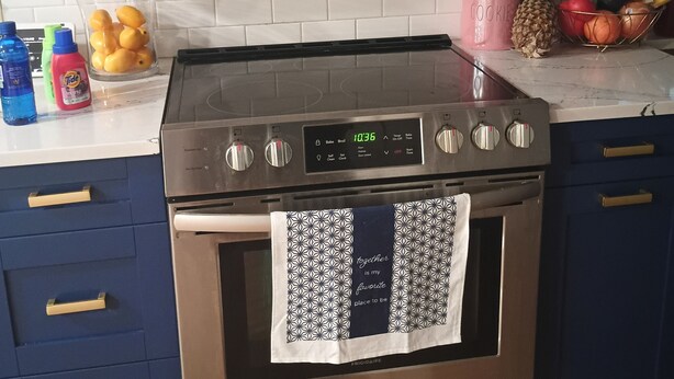 Cozinha com fogão de aço inoxidável, micro-ondas acima e painel de azulejos na parede. Há cestos de frutas no balcão e um recipiente rosa com a etiqueta 'COOKIE'.