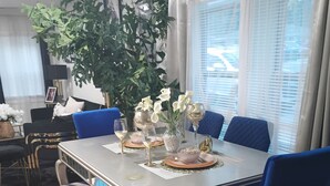Uma sala de jantar com mesa posta para quatro, cadeiras azuis, uma planta em vaso e uma janela com persianas.