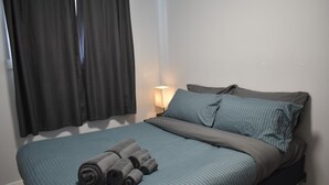 Een net opgemaakt bed met een blauwe spreideken en kussens, een opgerolde handdoek op het bed en een lamp op het nachtkastje.