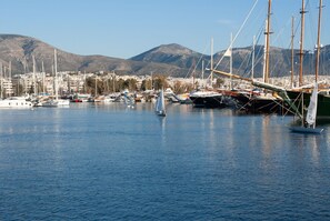 A marina with sailboats, a sailboat with a person on board, and a cityscape with mountains in the background. - Sun-Kissed Sailing Adventure in Islas Sarónicas (Alimos)
