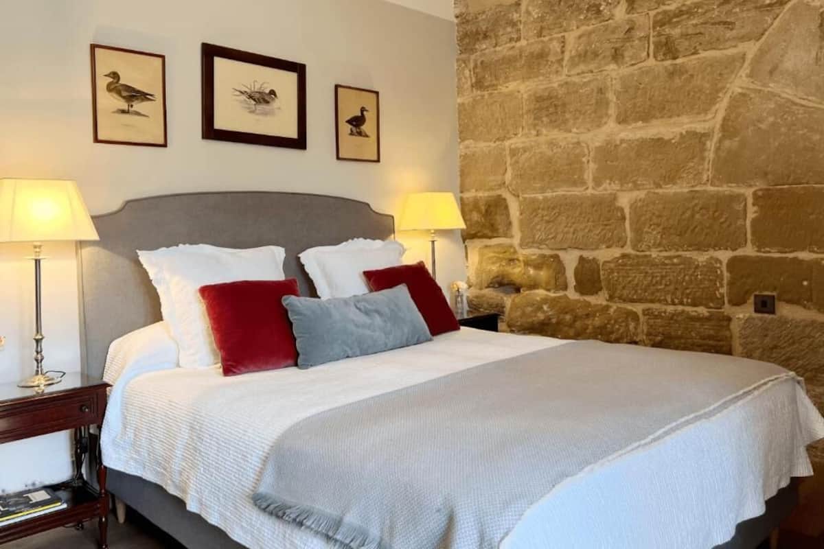 A bed with white and red pillows, a grey headboard, and a stone wall to the right.