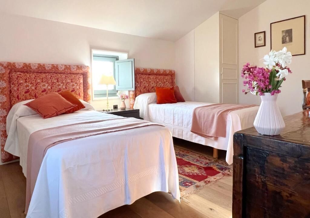 Una habitación de hotel con dos camas, piso de madera, un escritorio, una lámpara, un jarrón con flores y una ventana.