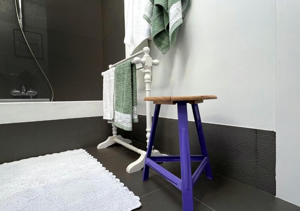 Un baño con una bañera blanca, un taburete de madera, un felpudo blanco y un tendedero de toallas con toallas verdes.