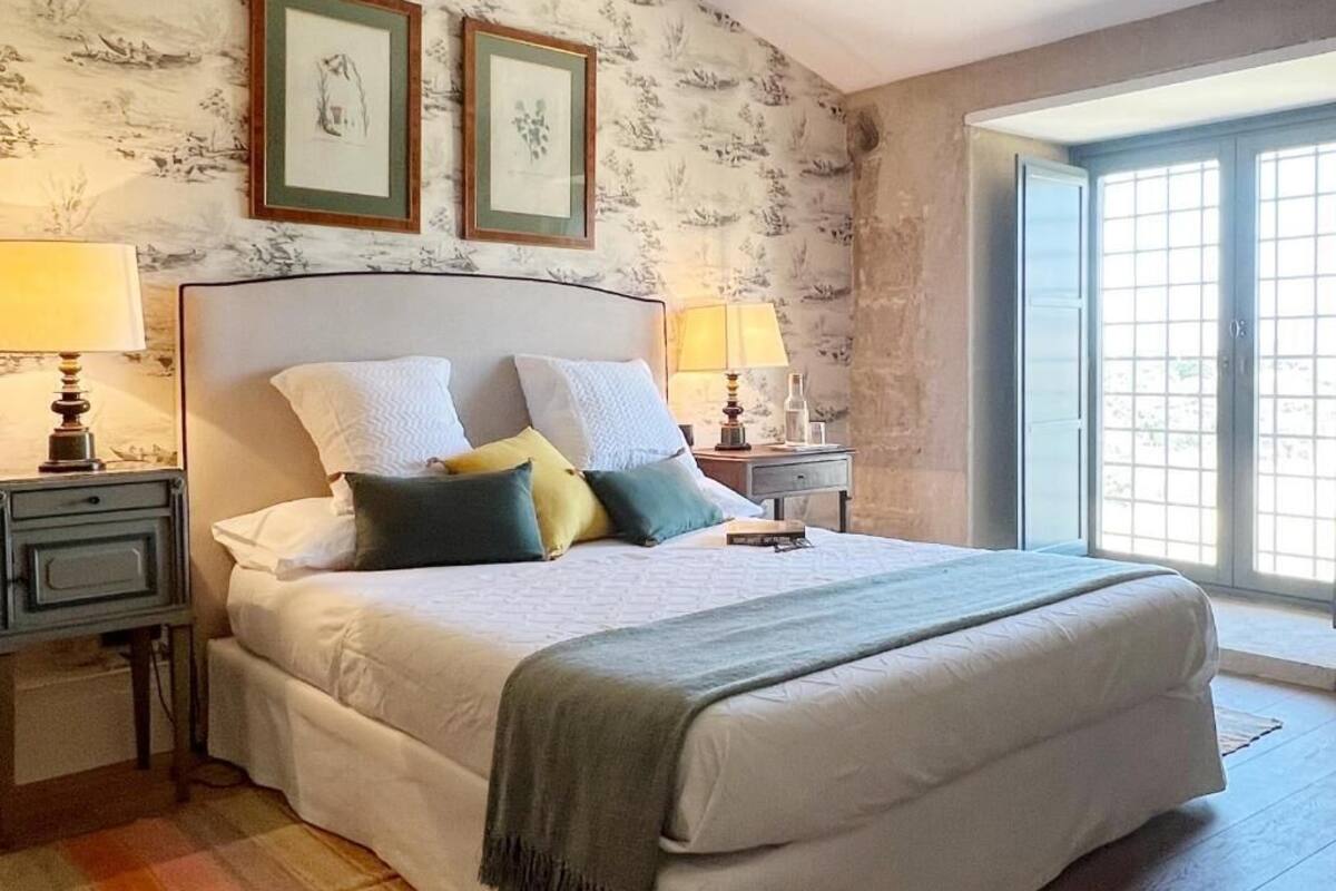 A bedroom with a large bed, two bedside tables with lamps, a window with a view, and floral wallpaper.