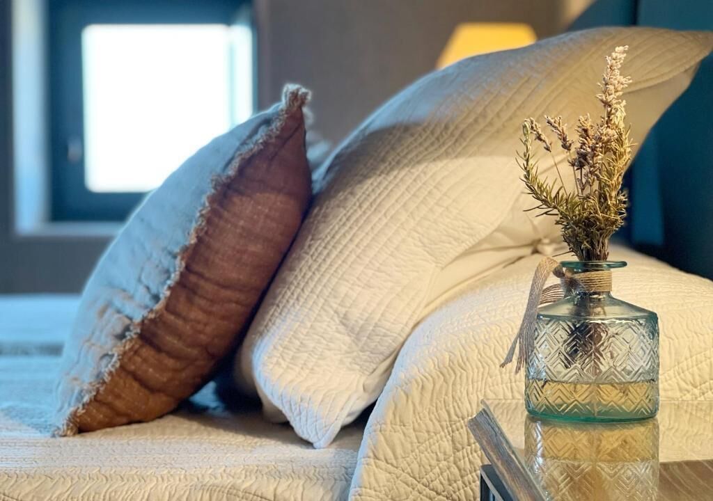 Una cama con un edredón texturizado y almohadas, un florero de vidrio con flores secas, y una ventana al fondo.