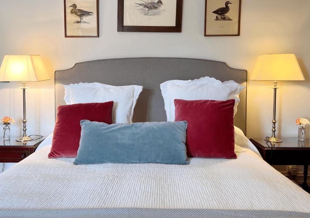A neatly made bed with white and red pillows, two lamps on either side, and framed artwork on the wall.