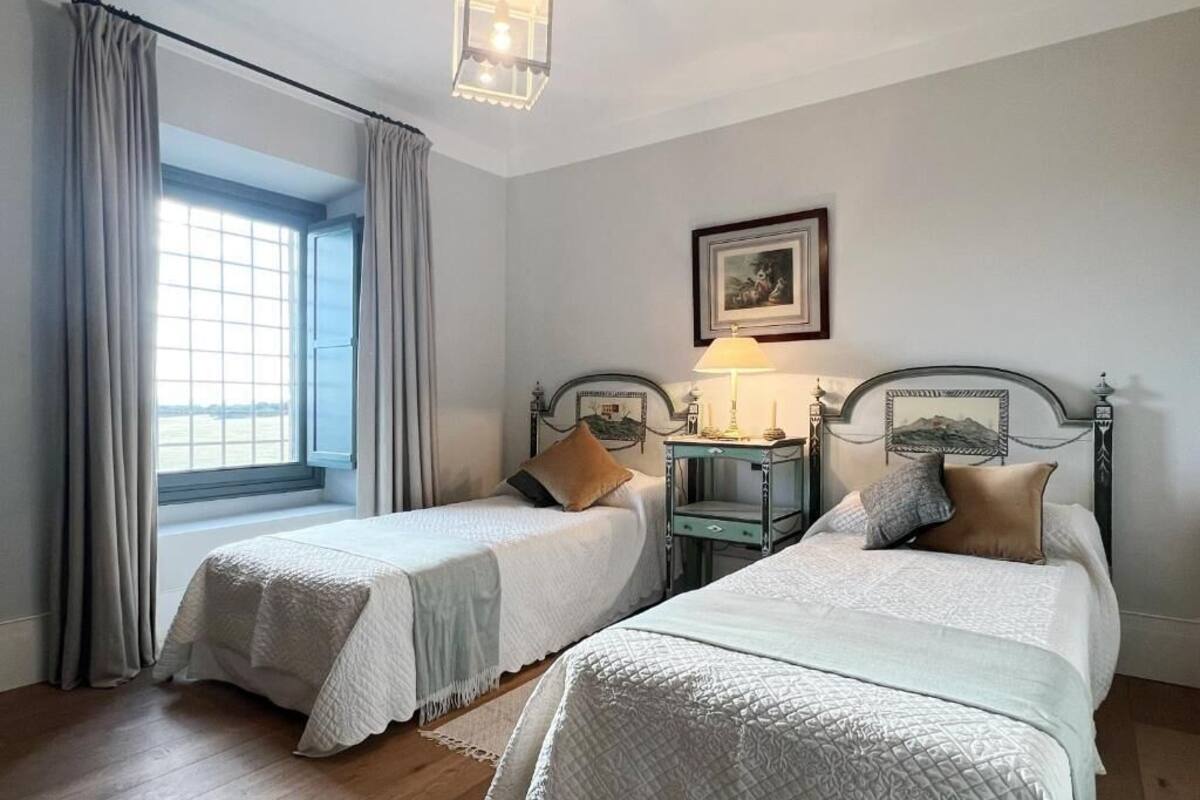 A room with two beds, a window with curtains, a bedside table with a lamp, and a framed picture on the wall.