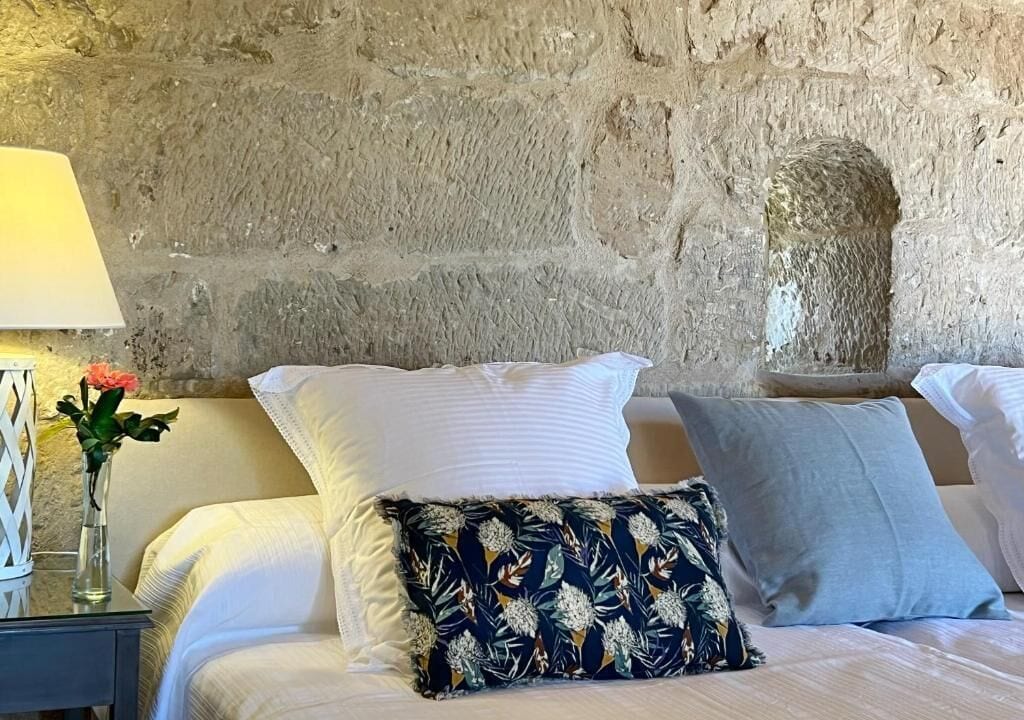 A bed with white and patterned pillows, a vase with a flower, and a lamp on a bedside table.