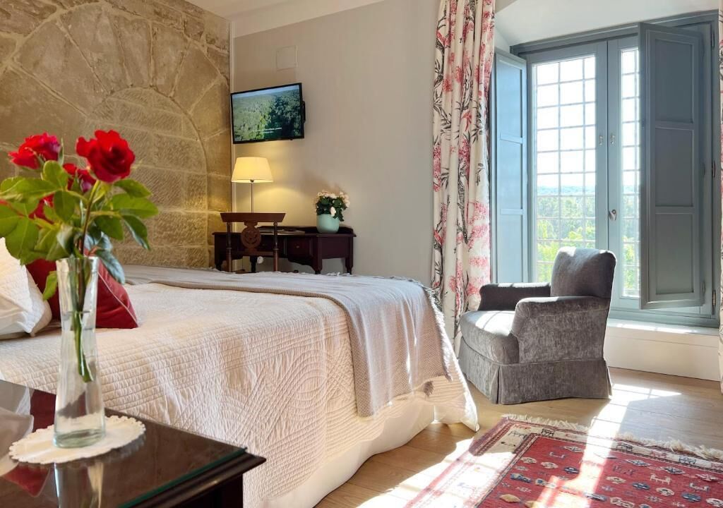 A bedroom with a bed, a desk, a chair, a vase with flowers, and a window with curtains.