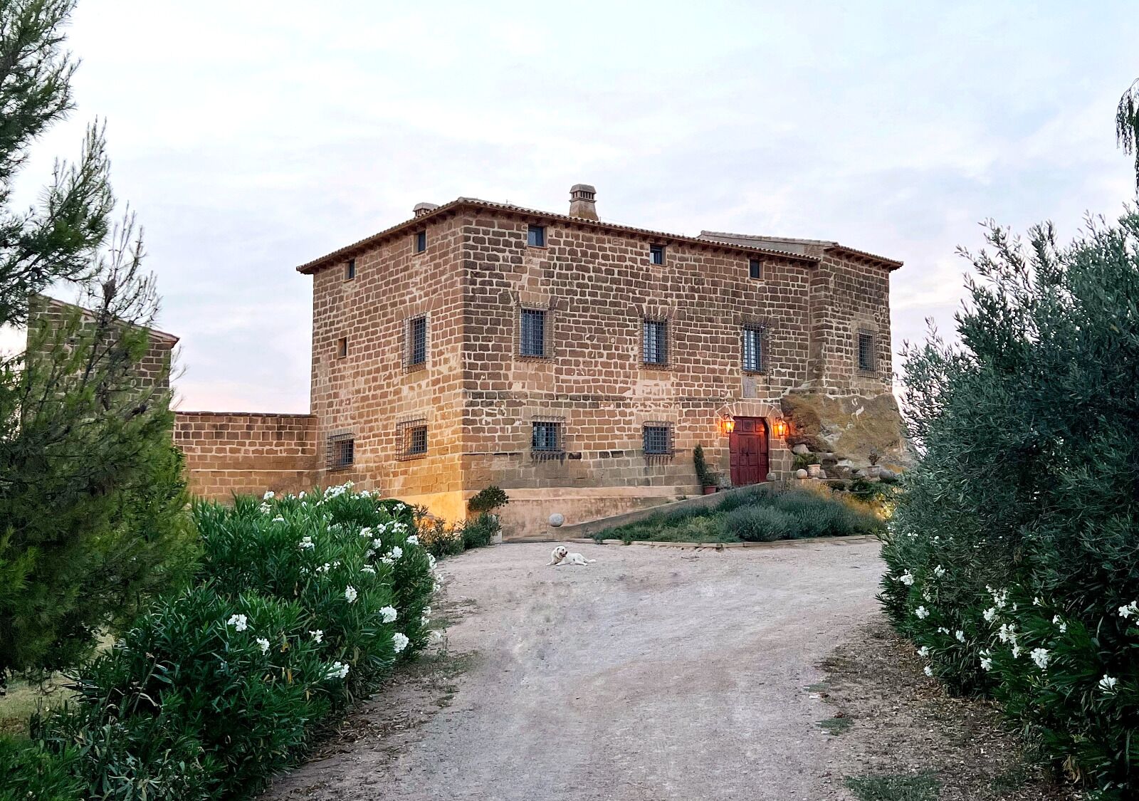 A stone building with a central entrance, surrounded by greenery and a gravel path.