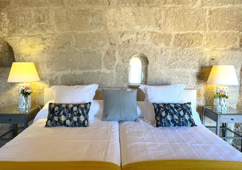 A double bed with a plain white bedspread, two pillows with a floral pattern, a single light blue pillow, and two bedside tables with lamps and vases.