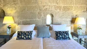 A double bed with a plain white bedspread, two pillows with a floral pattern, a single light blue pillow, and two bedside tables with lamps and vases.