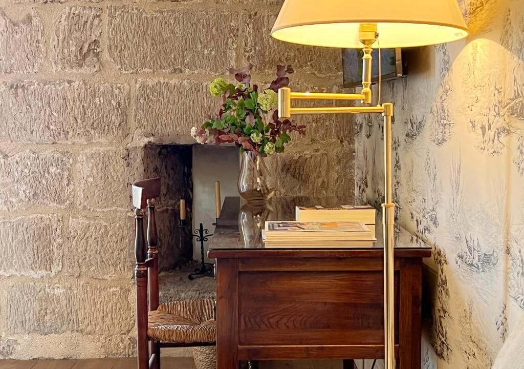 A wooden desk with a lamp, a vase with flowers, and a chair in a room with stone walls.