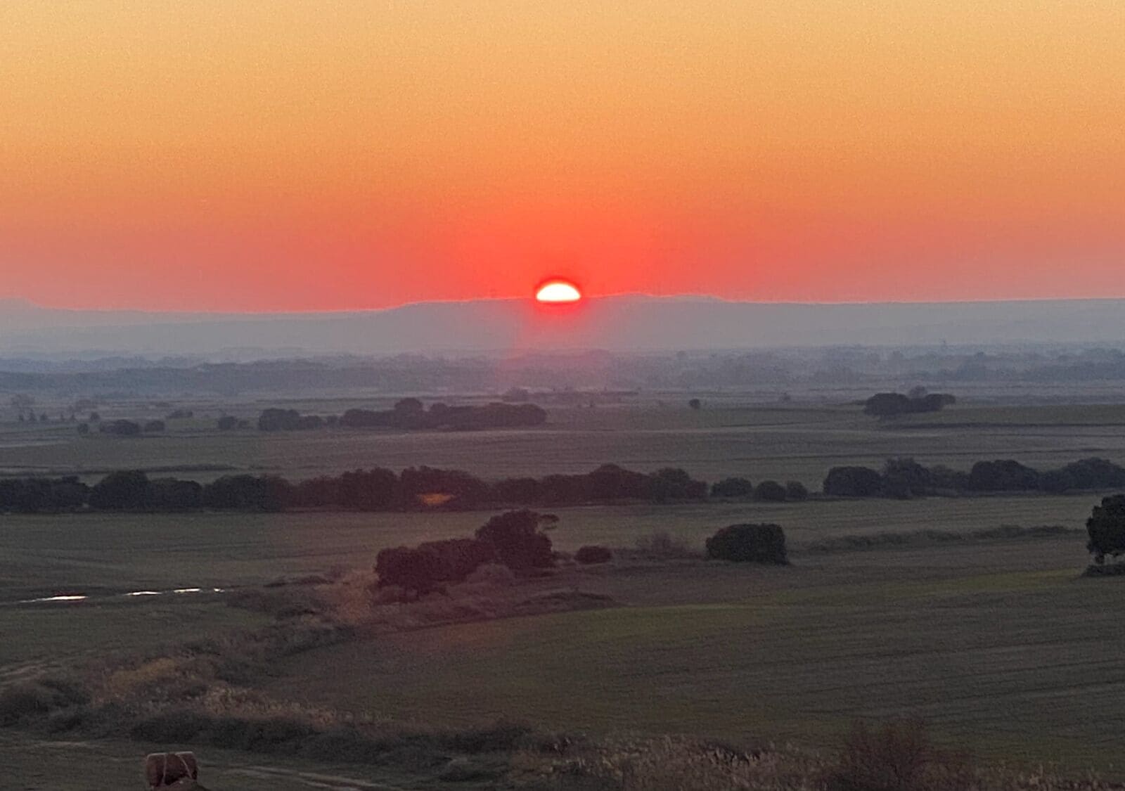 Un atardecer sobre un paisaje rural con campos y árboles dispersos.