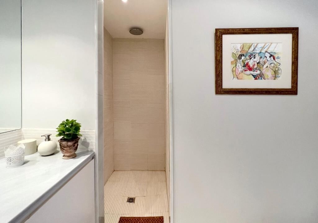 Un baño con encimera blanca, una planta en maceta, una obra de arte enmarcada en la pared y suelo de baldosas.