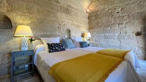 A bedroom with a stone wall, a bed with white bedding, two bedside tables with lamps, and a yellow throw blanket. - Castillo de Corvinos (Albero Alto)