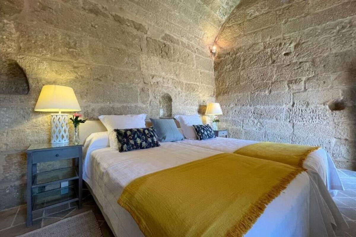 A bedroom with a stone wall, a bed with white bedding, two bedside tables with lamps, and a yellow throw blanket.