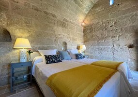 Un dormitorio con pared de piedra, una cama con ropa blanca, dos mesitas de noche con lámparas y una manta amarilla.