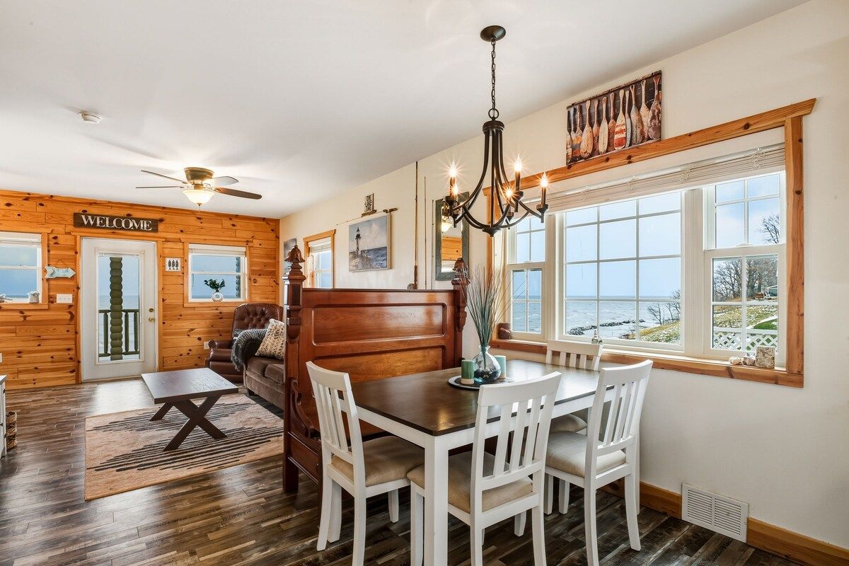 A wooden interior with a dining table, chairs, a sofa, a chandelier, and a view of the sea through large windows.