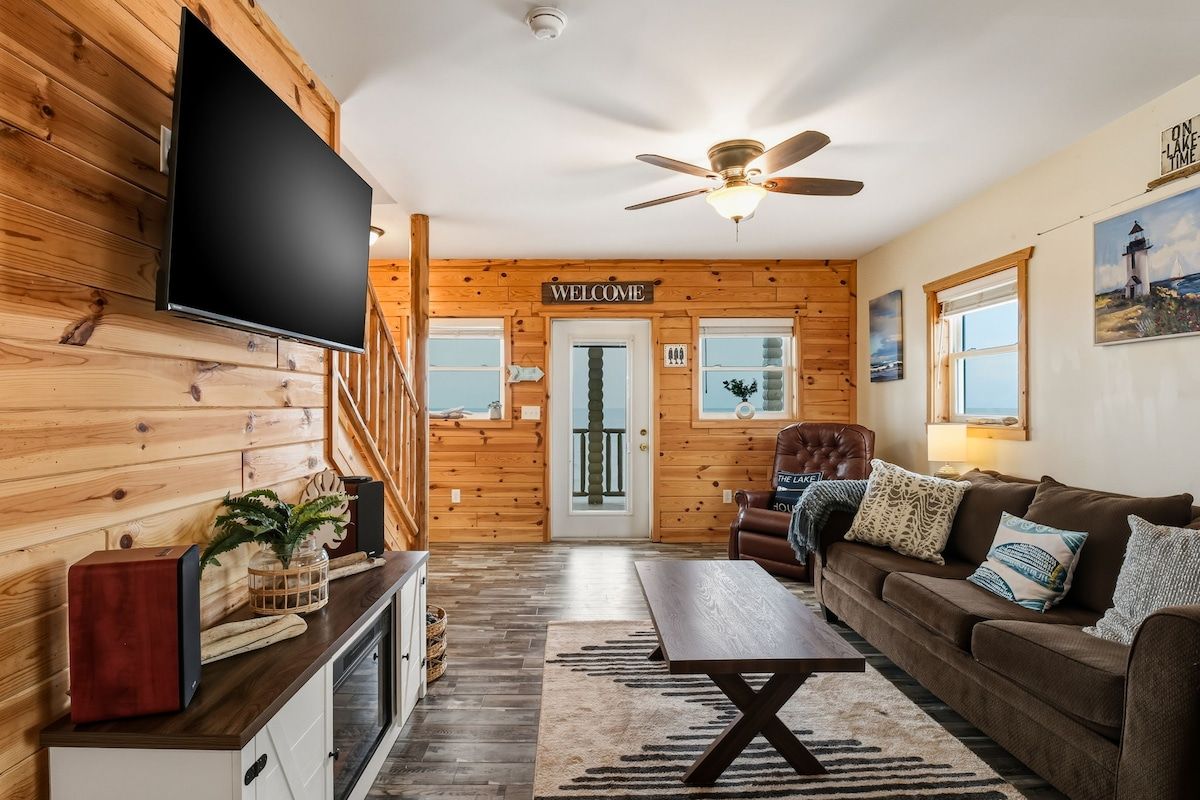 A wooden interior with a television mounted on the wall, a welcome sign, a ceiling fan, a couch, and a coffee table.