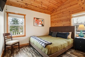 Una camera da letto con soffitto in legno, un letto con copripiumino verde, una sedia, un comodino con una lampada e una finestra vista alberi.