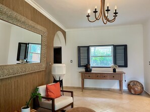 Ein Zimmer mit Holzpaneelen, einem großen Spiegel, einer Chandelierleuchtre, einem Fenster mit Fensterläden, einem Konsolentisch und einem Sessel mit Kissen.