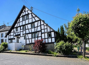 Une maison traditionnelle à colombages, avec un toit en pente, une cheminée bien visible et une rue pavée devant.