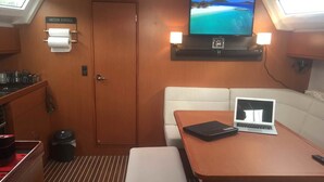 A compact galley with a wooden door, a small sink, and a toilet. A flat-screen TV is mounted above a wooden shelf, and a laptop is on a table. - Sun-Kissed Sailing Adventure in Islas Esporadas (Skiathos)