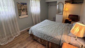 Um quarto com cama, abajur, mesa de cabeceira de madeira, espelho, janela com cortinas e prateleira na parede.