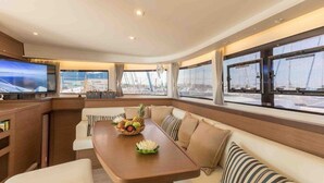 A modern interior with a dining area, cushioned seating, a television, and large windows offering views of the sea. - Sun-Kissed Sailing Adventure in the Stunning Islas Cícladas (Paros)