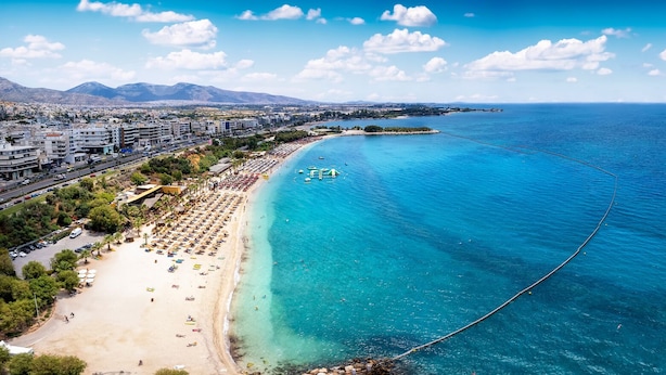 Paisagem costeira com praia, águas azuis cristalinas e uma cadeia de montanhas ao longe.