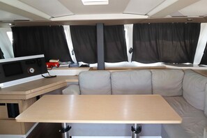 A compact RV interior with a central seating area, a small kitchenette, and a window with curtains. - Sun-Kissed Sailing Adventure in Islas Sarónicas (Alimos)