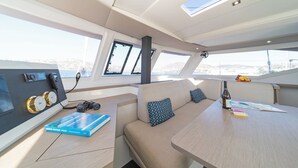 Interior de un yate con panel de control, zona de estar y una mesa con una botella y documentos.