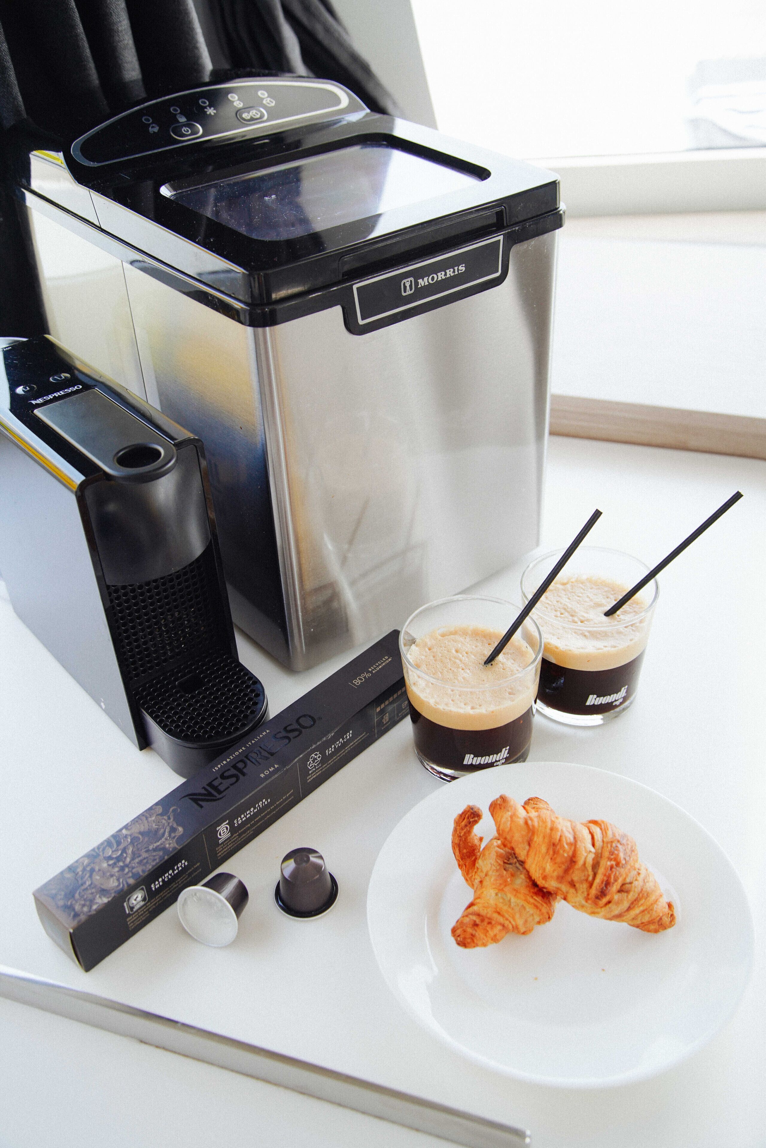A coffee maker, two glasses of coffee, a croissant, and a coffee pod box.