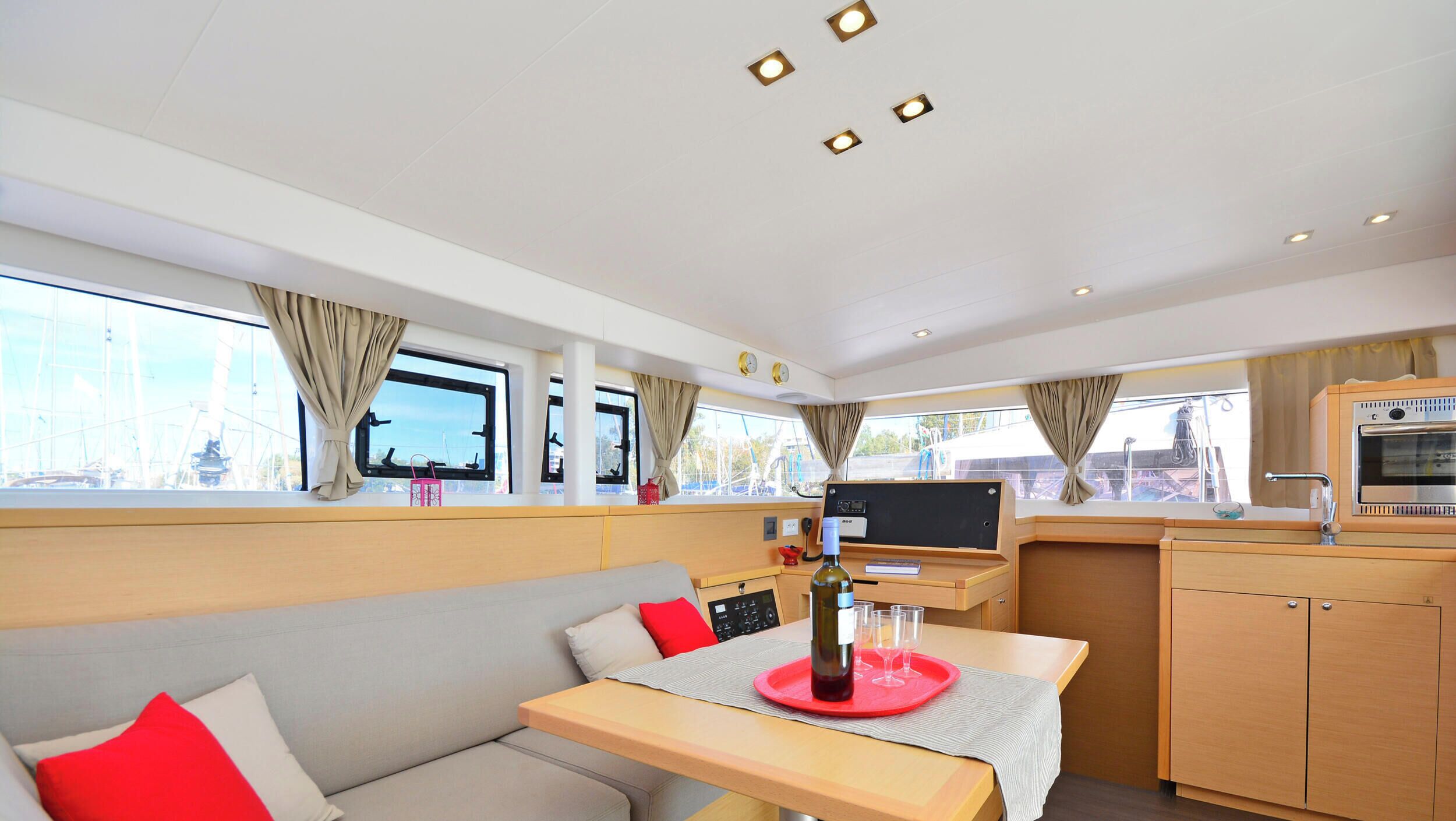 A compact, well-lit room with a sofa, a dining table with a bottle and glasses, a small kitchen area with an oven, and large windows with curtains.