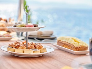 A tiered dessert stand with macarons and pastries, a plate of scones, and a slice of cake with yellow frosting on a table.