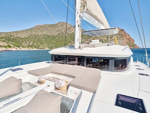A sailboat with a deck area featuring cushioned seating, a small table with a basket, and a view of the sea and a rocky coastline.