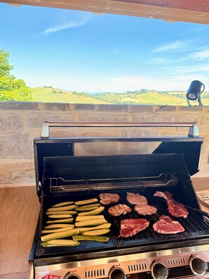 Ein Grill mit Fleisch und Zucchini auf dem Rost, vor einer Steinmauer mit Blick auf grüne Felder.