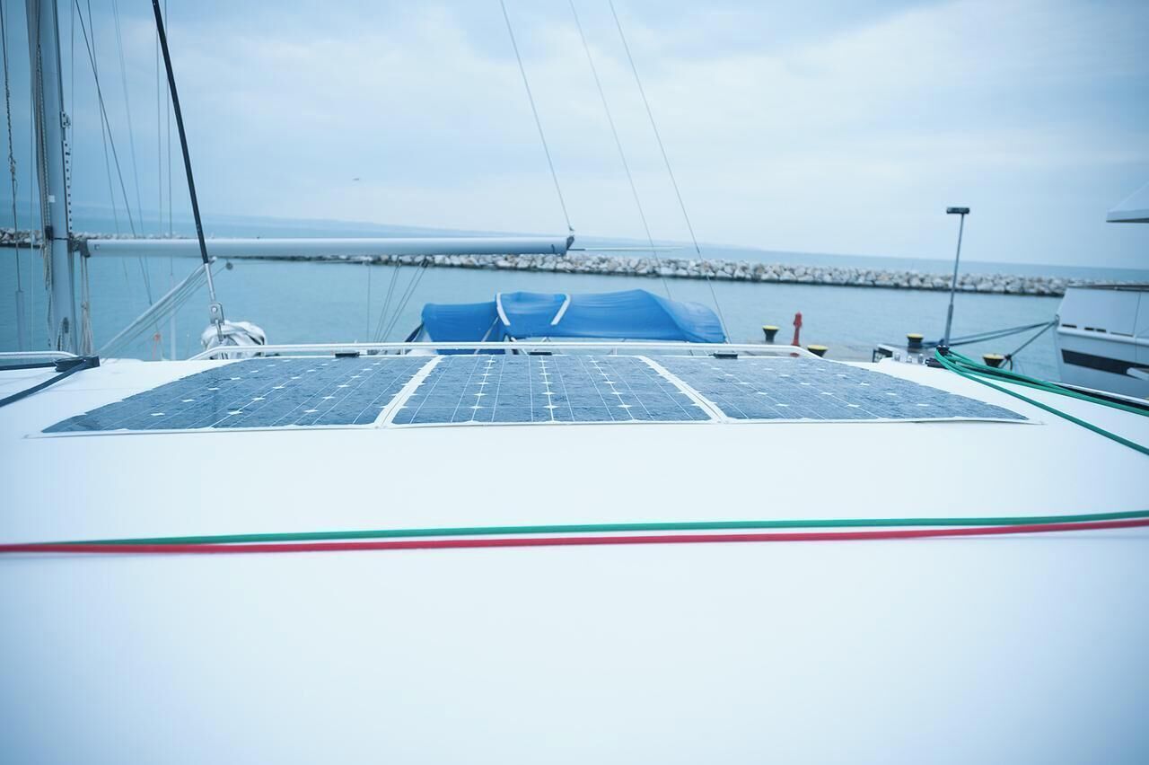 Ein Segelboot mit Solarmodulen auf dem Deck, einer blauen Plane und Blick auf Wasser und Ufer.