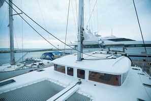 Ein Segelboot liegt in einer Marina festgemacht, mit mehreren Masten und Segeln, einem überdachten Bereich mit Sitzgelegenheiten und Blick auf das Wasser.