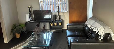 Una sala de estar con una mesa de centro de vidrio, un sofá de cuero, un televisor y una ventana con persianas.