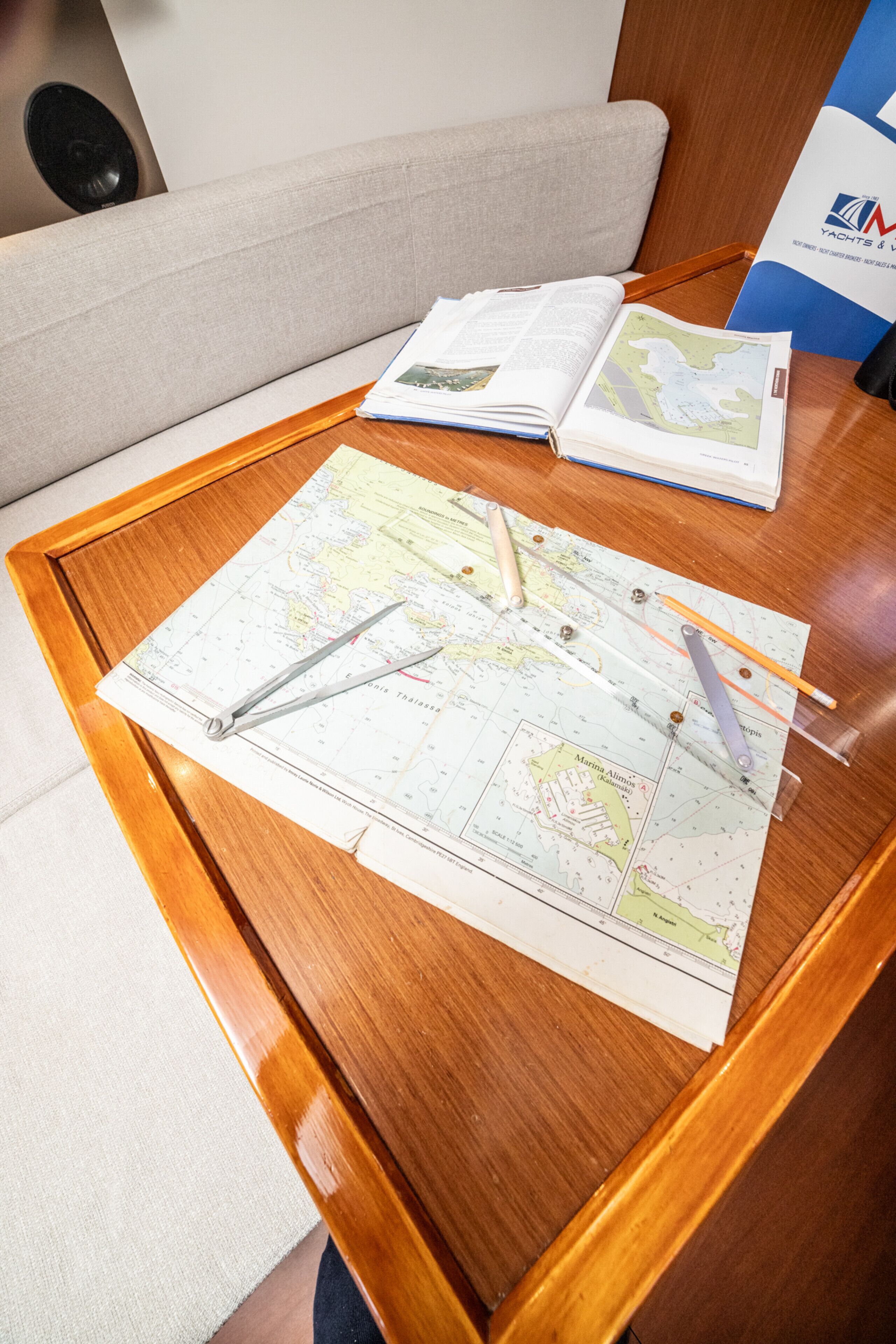 A wooden table with a map, compass, and pencil, a sofa, and a brochure in a room with a wooden floor and white walls.