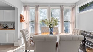 Een eetgedeelte met een houten tafel, beiges bekleden stoelen, een vaas met bloemen en grote ramen met gordijnen.