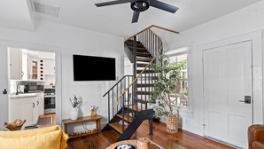 A modern living room with a wooden floor, a round coffee table, a sofa, a TV mounted on the wall, a staircase with a black railing, and a door leading to the outside. - Historic Wilmington Estate, 5BR/3 Suites - Riverwalk, VIP Parking (Wilmington)