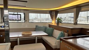 A cabin with a table, bench, and seating area, featuring a view of the marina through large windows. - Sun-Kissed Sailing Adventure in Islas Sarónicas (Alimos)