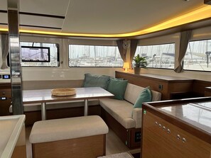 A cabin with a table, bench, and seating area, featuring a view of the marina through large windows. - Sun-Kissed Sailing Adventure in Islas Sarónicas (Alimos)