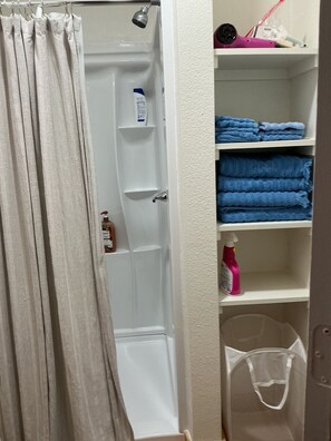 A bathroom with a shower curtain, a white bathtub, shelves with folded towels, and a trash bin. - Adorable 1-bedroom barn in Mineola, East Texas also known as Lake Country (Mineola)