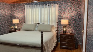 A bedroom with a wooden ceiling, floral wallpaper, a bed with a wooden frame, two bedside tables with lamps, and a window with curtains. - Adorable 1-bedroom barn in Mineola, East Texas also known as Lake Country (Mineola)