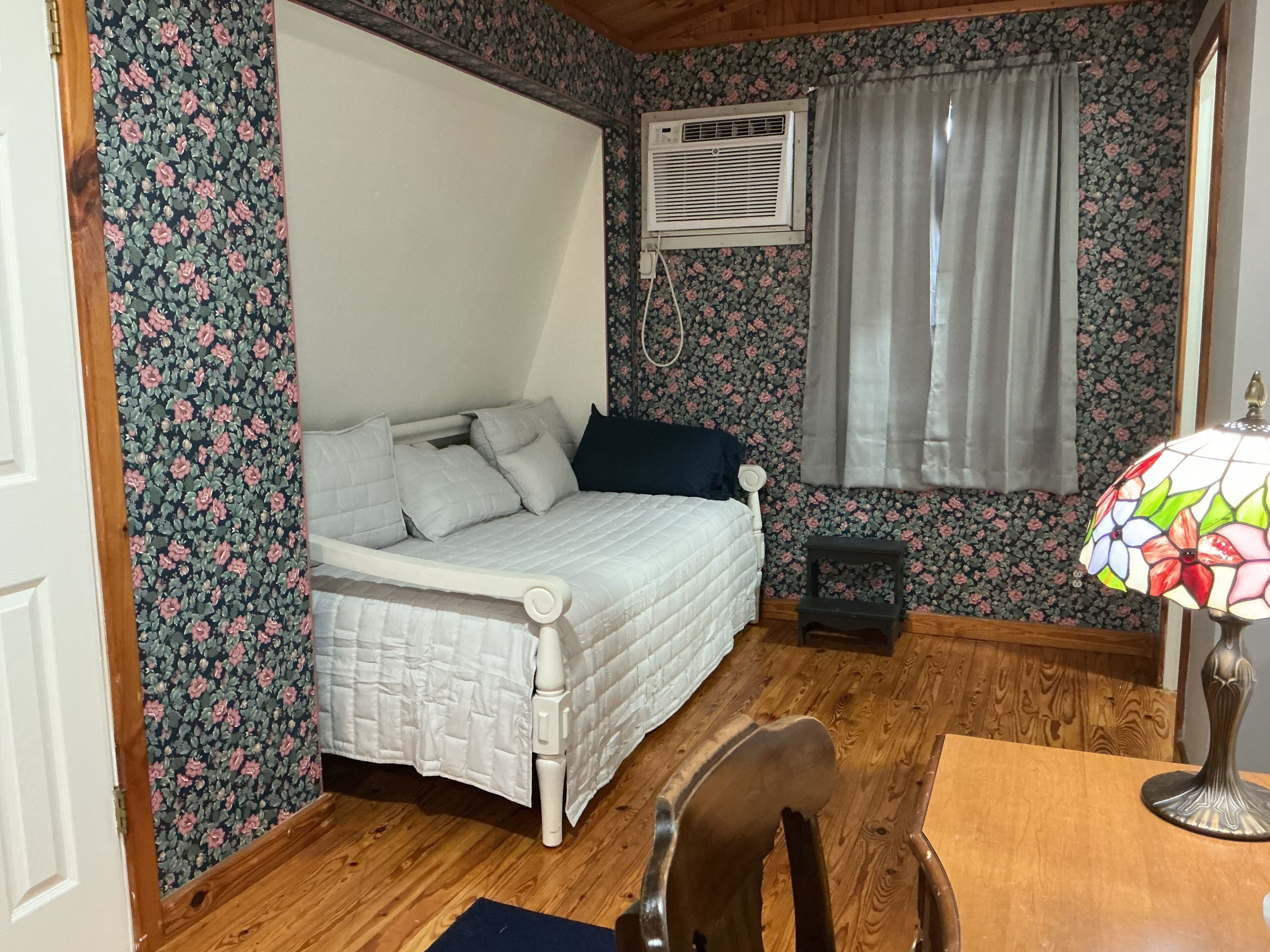 Une chambre à coucher comprenant un lit, une petite table, une chaise, une fenêtre avec des rideaux, un appareil de climatisation et un papier peint à motifs floraux.